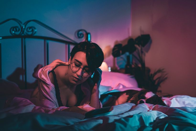 A woman with glasses relaxes in a cozy, dimly lit bedroom setting.